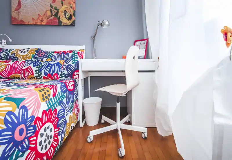 Bright, colorful bedroom with a patterned bedspread, a small white desk and a rolling chair by a sunlit window — cozy and well-styled for a rental listing.