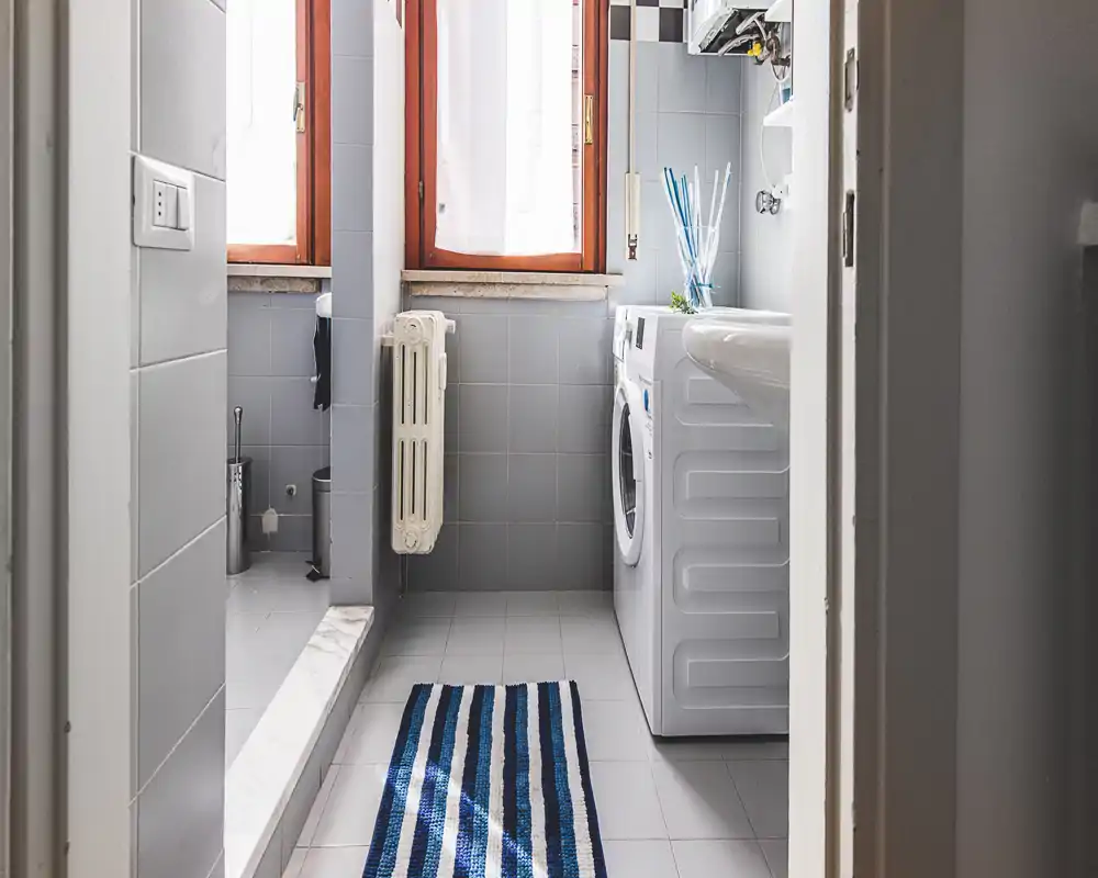 Compact bathroom with washing machine and sink in a bright tiled space, featuring a striped floor mat and natural light from the window.