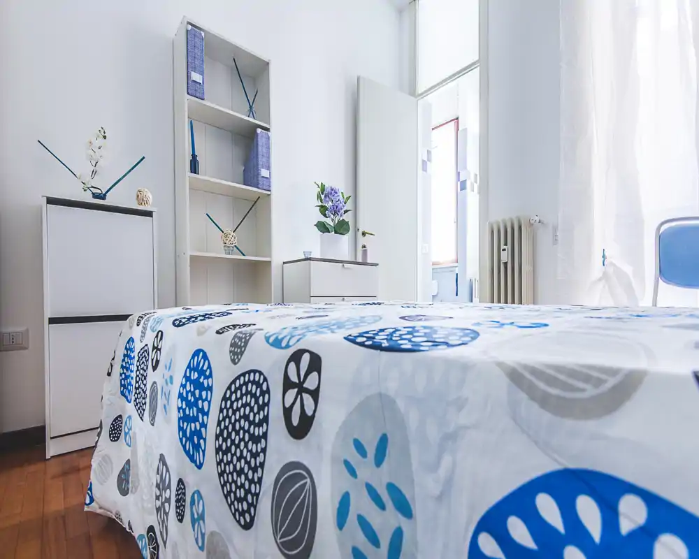Bright, modern bedroom with patterned blue-and-gray bedding, simple shelving and a small bedside cabinet; abundant natural light and minimal, tidy decor.