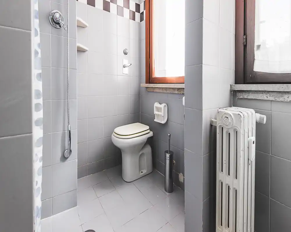 Compact tiled bathroom with a toilet, hand shower, window and radiator; neutral gray tiles and natural light make the space practical and bright.