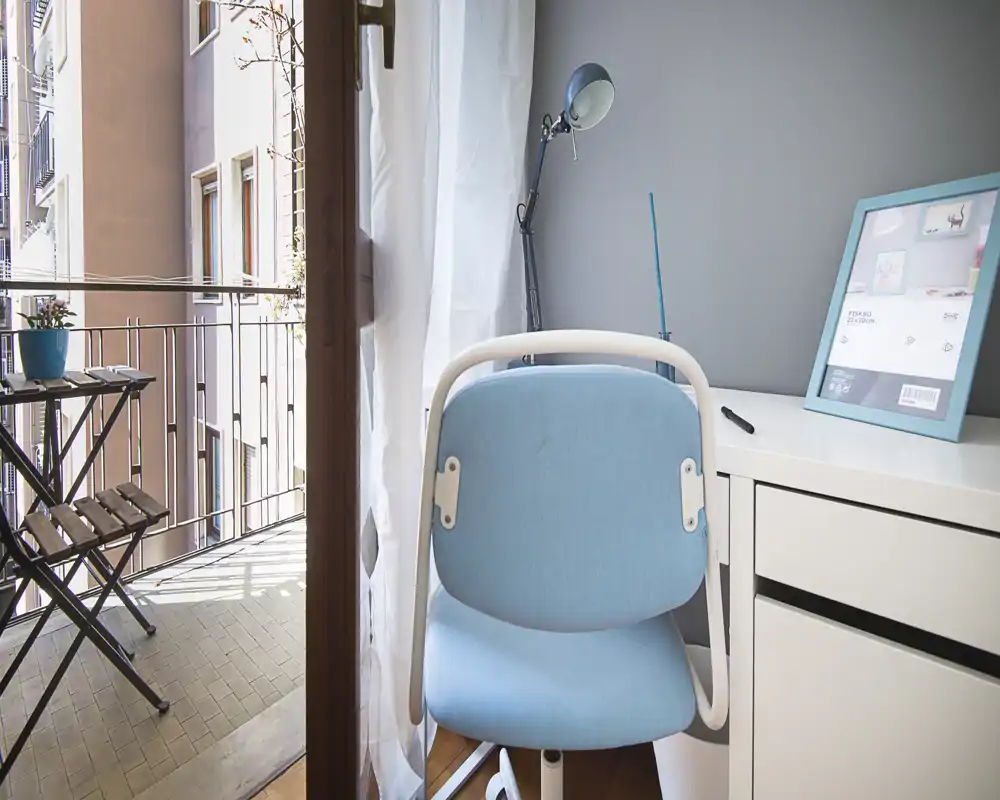 Bright home office corner opening onto a small balcony. The compact desk and blue chair create a tidy workspace with natural light and a pleasant outdoor view.