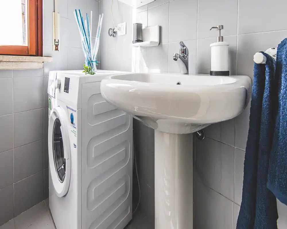 Compact bathroom corner featuring a pedestal sink and a front-loading washing machine under a bright window, with tidy accessories and blue towels.