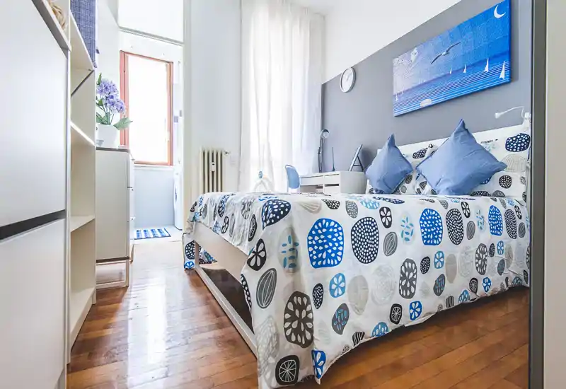Bright, modern bedroom with a double bed dressed in patterned linens, decorative pillows and a small desk by the headboard. Natural light from a window creates a welcoming, well-styled space ideal for listings.