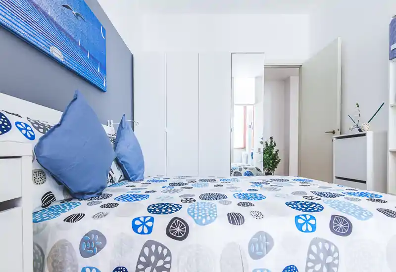 Bright, modern bedroom with a patterned blue-and-grey bedspread, matching cushions and clean white furniture. The composition highlights the bed and a built-in wardrobe, creating a fresh, coastal feel.