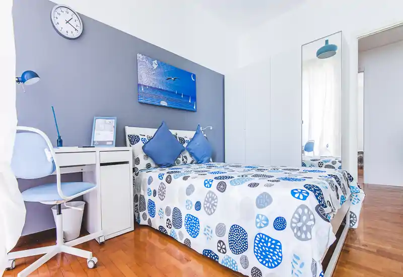 Bright, neatly staged bedroom with a double bed dressed in blue-and-white patterned linens, coordinating cushions and a small desk for work. Clean contemporary styling, hardwood floors and a full-height mirror add space and light.