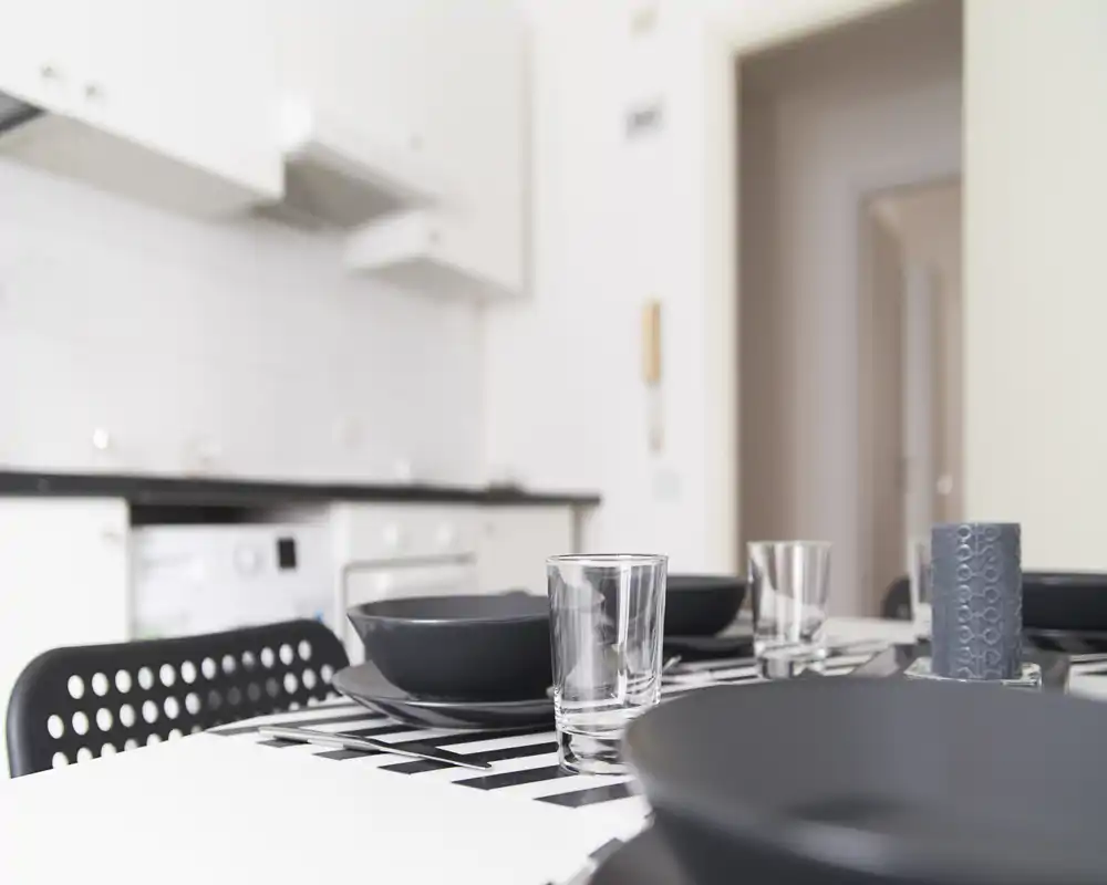Contemporary dining area with a neatly set table: black plates, clear glasses and a decorative candle on a striped runner, adjacent to a compact white kitchen.
