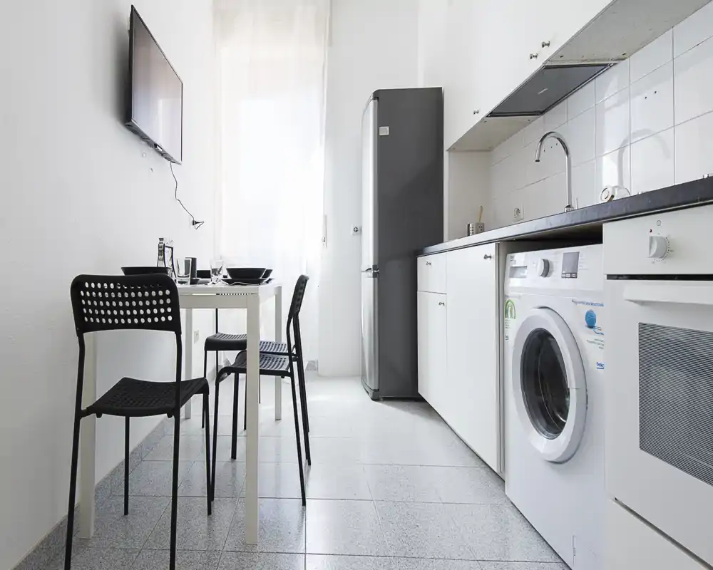 Bright modern kitchenette with white cabinets, integrated appliances and a small dining table; clean minimalist styling and natural light from a window.