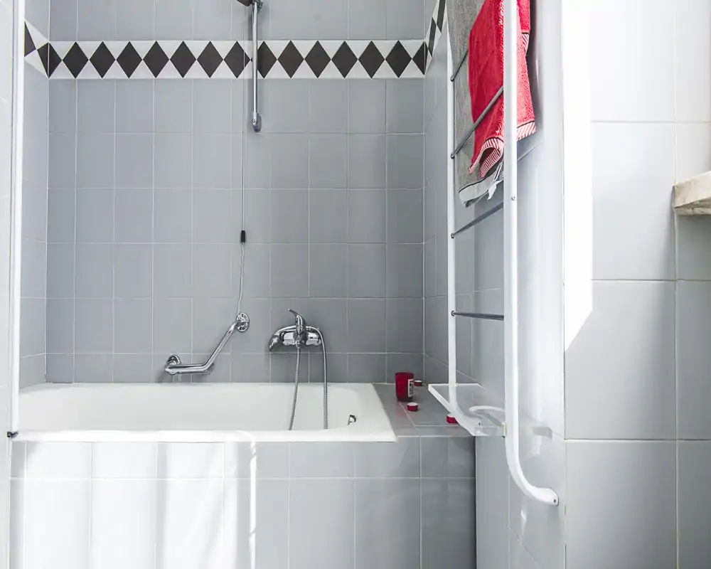 Clean, light bathroom with a built-in bathtub, tiled walls and chrome fixtures. Natural light brightens the space and a heated towel rail with red towels adds a touch of color.