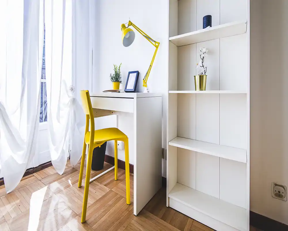 Bright corner workspace in a living area with a compact white desk, yellow chair and lamp; airy window curtains and a tall shelving unit add storage and light.