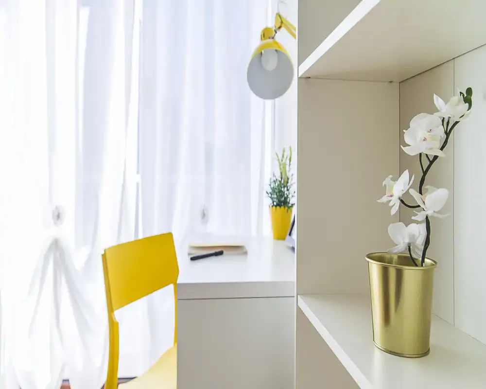 Bright, minimal workspace corner with a white desk, yellow chair and matching lamp, complemented by small plants and a decorative orchid.