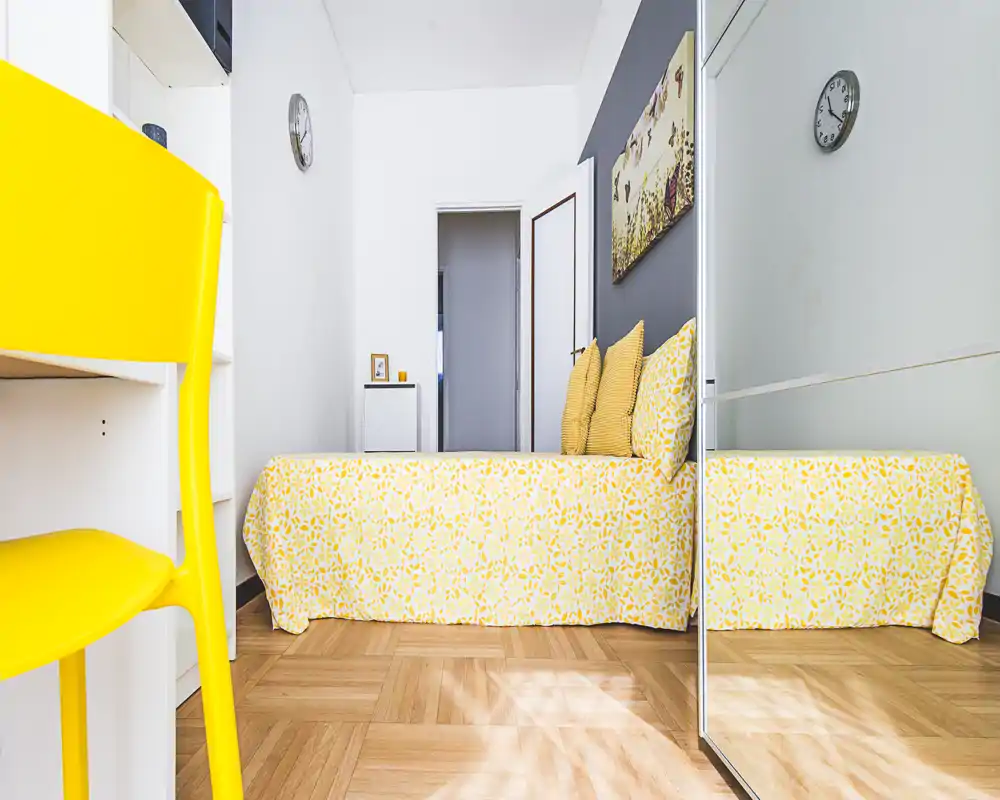 Bright, compact bedroom with a patterned yellow bedspread, matching cushions and a full-height mirrored wardrobe that visually enlarges the space.