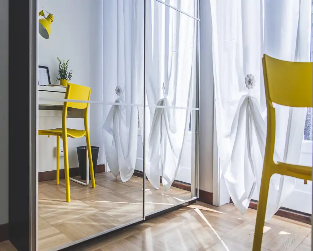 Bright bedroom corner with mirrored wardrobe reflecting white curtains and a yellow chair beside a small desk; wooden parquet and natural light create a fresh, minimal atmosphere.