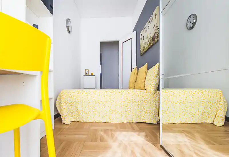 Bright, compact bedroom with a patterned yellow bedspread, matching cushions and a full-height mirrored wardrobe that visually enlarges the space.