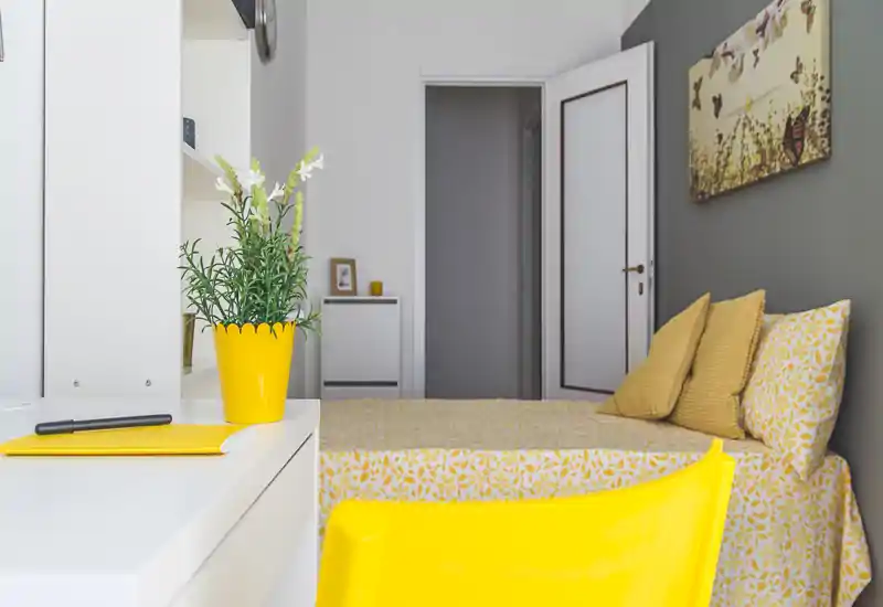 Bright, stylish bedroom with coordinated yellow accents — patterned bedding, cushions and a matching chair create a warm, modern atmosphere. A tidy desk with a yellow plant pot in the foreground adds useful staging for a rental listing.