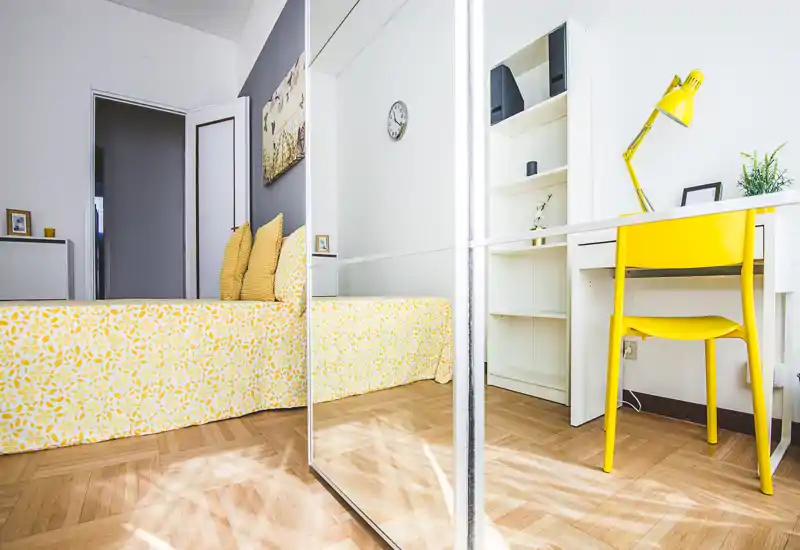 Bright, well-staged bedroom with a yellow patterned bedspread, matching cushions and a compact desk area; mirrored wardrobe adds depth and natural light.