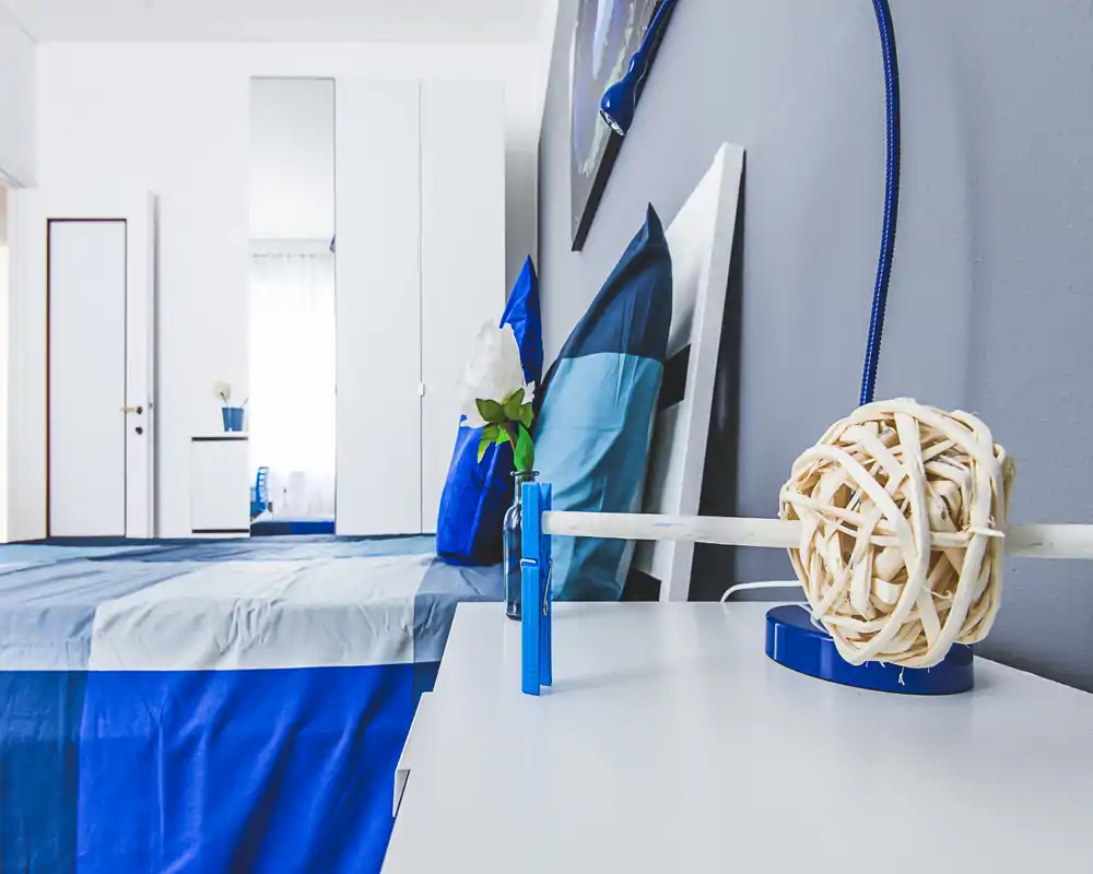 Bright, modern bedroom with blue bedding, decorative pillows and a bedside table styled with a lamp and vase. The room feels fresh and well lit with a wardrobe and mirror in the background.