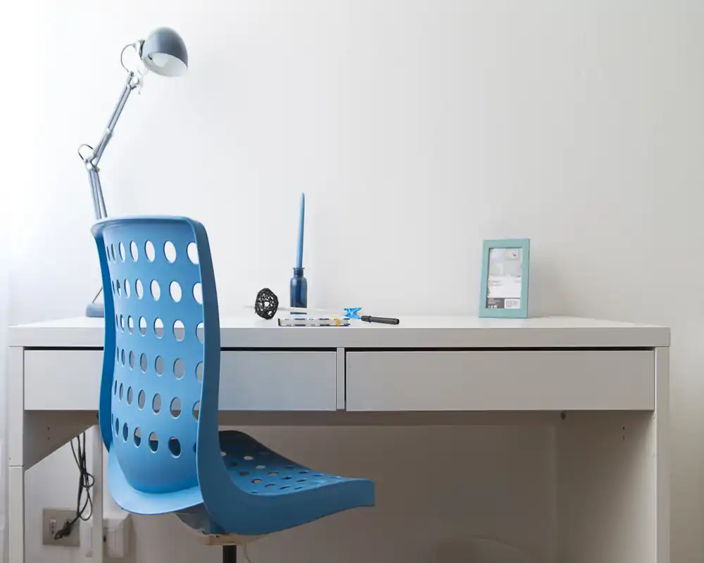 Minimal study corner with a white desk and a modern blue perforated chair, styled with a desk lamp and a few small decorative objects — bright, clean and functional workspace.