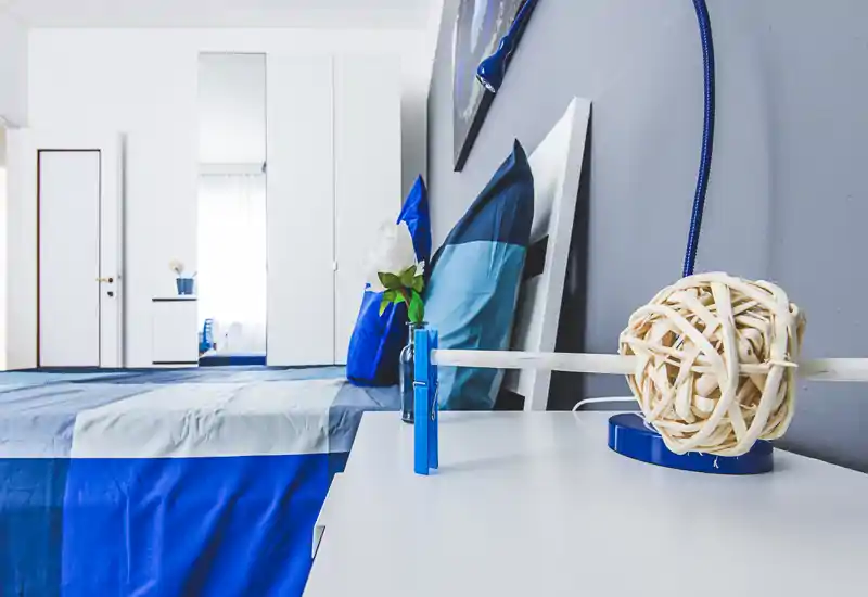 Bright, modern bedroom with blue bedding, decorative pillows and a bedside table styled with a lamp and vase. The room feels fresh and well lit with a wardrobe and mirror in the background.