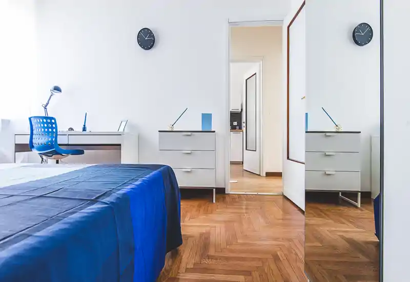 Bright, modern bedroom with blue bedding, a desk and chair by the window, parquet flooring and a mirrored wardrobe reflecting the room.