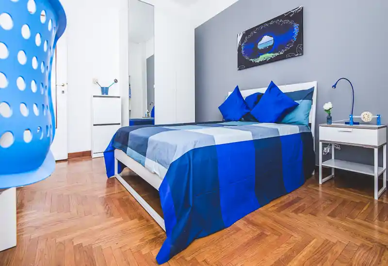 Bright, modern bedroom with a neatly made double bed in blue tones, bedside table with lamp and stylish wall art—ideal for rental photos highlighting comfort and color-coordinated decor.