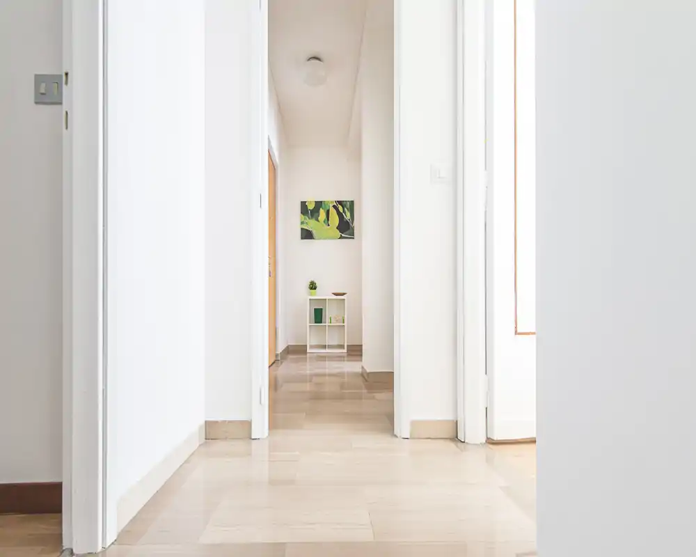Ingresso/corridoio luminoso e minimale con pavimento in legno chiaro, pareti bianche e un piccolo mobiletto sul fondo; composizione pulita e simmetrica, ideale per presentare l’ingresso dell’appartamento.