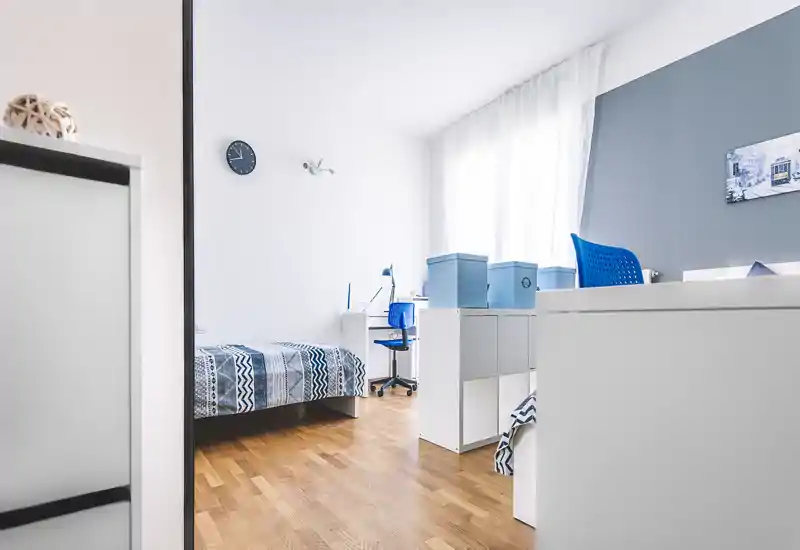 Camera da letto moderna e luminosa con pavimento in legno, letto singolo con copriletto a pattern e angolo studio con sedia blu; finestra ampia fornisce molta luce naturale.