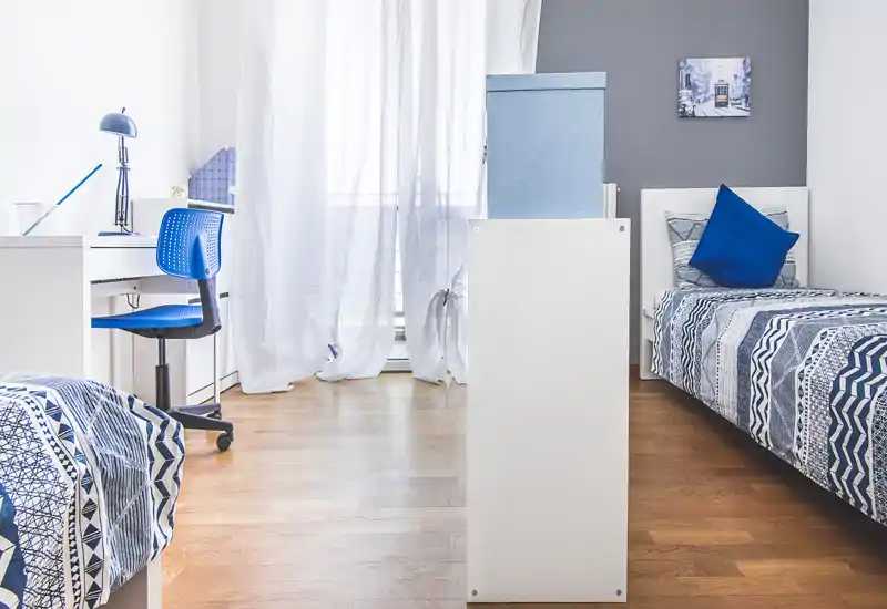 Camera luminosa e ordinata con due letti singoli, tessili coordinati blu e una scrivania vicino alla finestra — ideale per studenti o affitti condivisi.