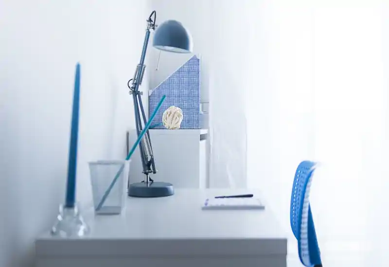 Angolo giorno/studio luminoso e minimal con scrivania bianca, sedia blu e lampada regolabile — ambiente pulito e arioso ideale per concentrarsi.