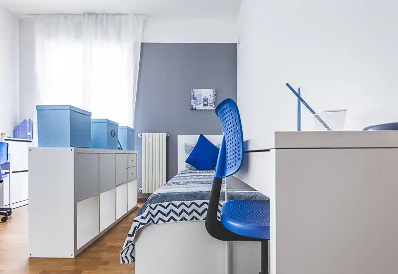 Camera singola luminosa e moderna con accenti blu coordinati, letto comodo con biancheria a motivo, scrivania e contenitori—ideale per studenti o affitti brevi.