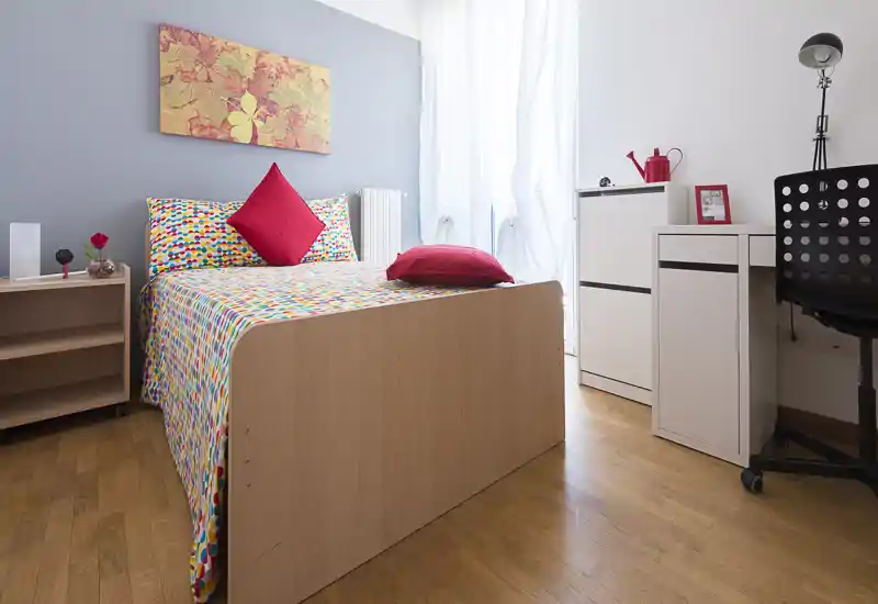 Camera da letto singola luminosa e ordinata con letto in legno, biancheria colorata e cuscini rossi, piccolo scrittoio e luce naturale da una grande finestra.