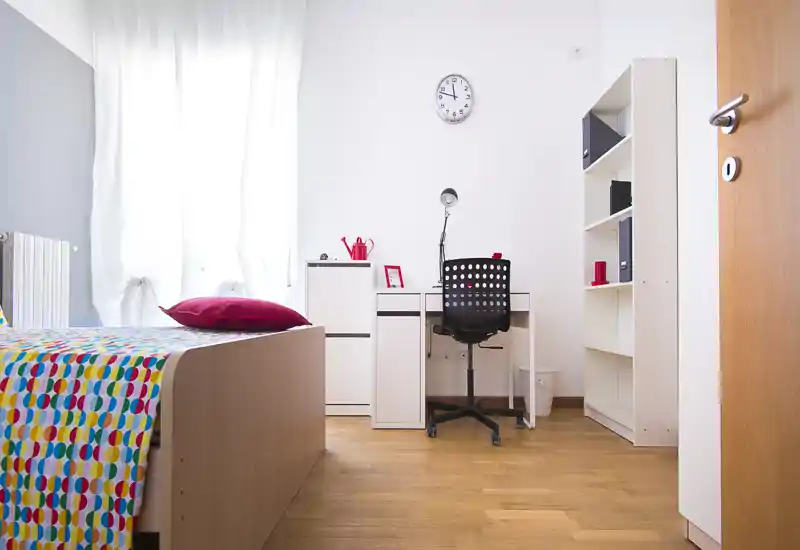 Camera singola luminosa e ordinata con copriletto colorato, scrivania e scaffale — ideale per uno studente o un professionista. La luce naturale dalla grande finestra crea un ambiente fresco e accogliente.