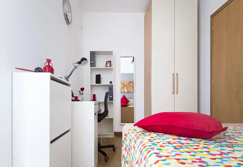 Camera singola luminosa e moderna con copriletto colorato e cuscino rosso. La stanza dispone di scrivania con scaffali, armadio a tutta altezza e specchio a figura intera — perfetta per uno studente o un giovane professionista.