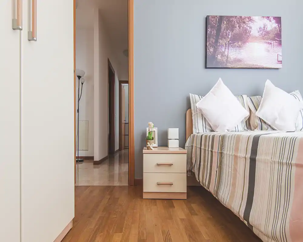 Rincón de dormitorio ordenado y bien iluminado con cama de rayas, cojines decorativos y pequeña mesita de noche—ideal para un anuncio acogedor.