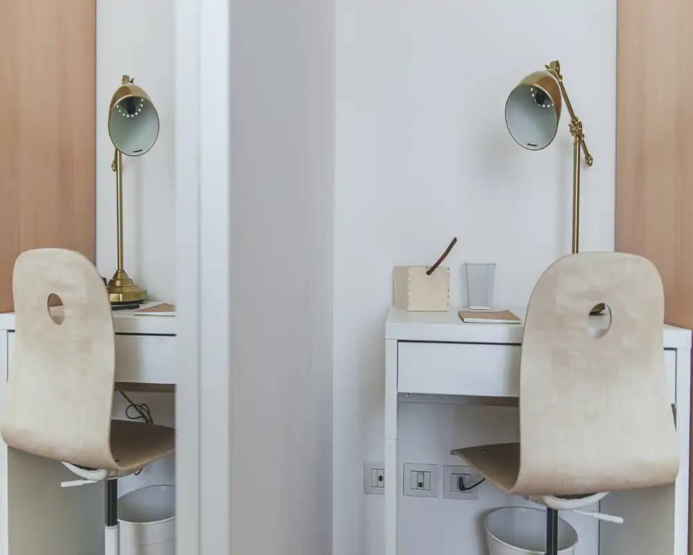 Pequeño espacio de trabajo luminoso con dos sillas en madera clara y escritorios blancos. Lámparas doradas y accesorios mínimos crean un rincón de estudio moderno y limpio.