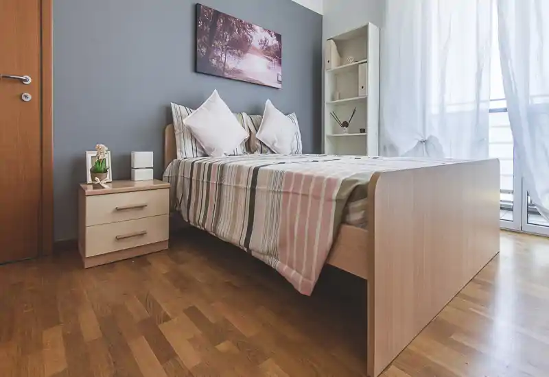 Dormitorio luminoso y ordenado con cama doble, mesita, estantería y luz natural suave a través de cortinas translúcidas — perfecto para anuncios de alquiler que destacan confort y sencillez moderna.