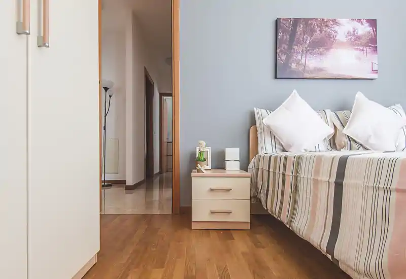 Rincón de dormitorio ordenado y bien iluminado con cama de rayas, cojines decorativos y pequeña mesita de noche—ideal para un anuncio acogedor.