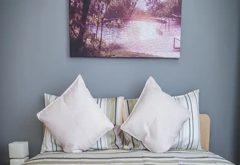 Imagen luminosa de dormitorio con cama bien hecha, dos grandes cojines decorativos, ropa de cama a rayas y un cuadro sobre la cabecera — perfecta para un anuncio acogedor.