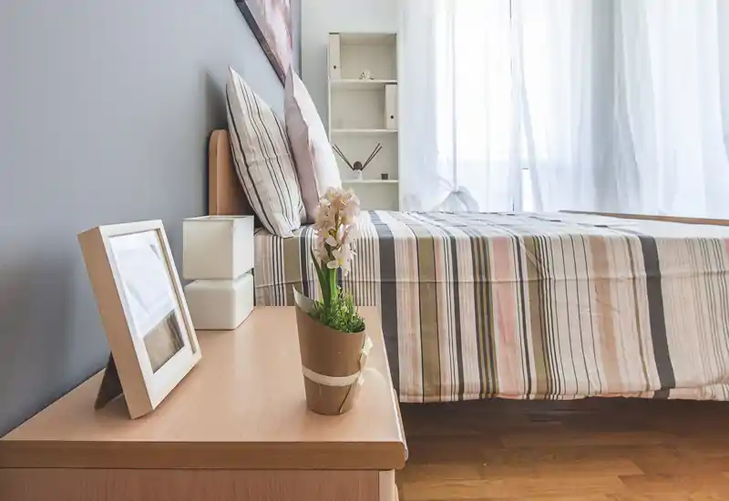 Dormitorio luminoso y ordenado con cama de rayas, mesita de noche y luz natural suave desde una ventana con cortina transparente — acogedor y listo para el anuncio.