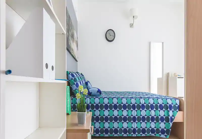 Camera da letto luminosa e moderna con copriletto a motivo blu-verde, cuscini coordinati e arredi minimalisti. L'immagine mette in evidenza il letto, la sistemazione è ordinata e la luce naturale è buona.