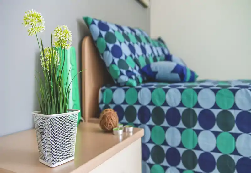 Angolo camera luminoso e curato con biancheria a motivo geometrico turchese e blu, cuscini coordinati e un comodino con decorazioni e una pianta.