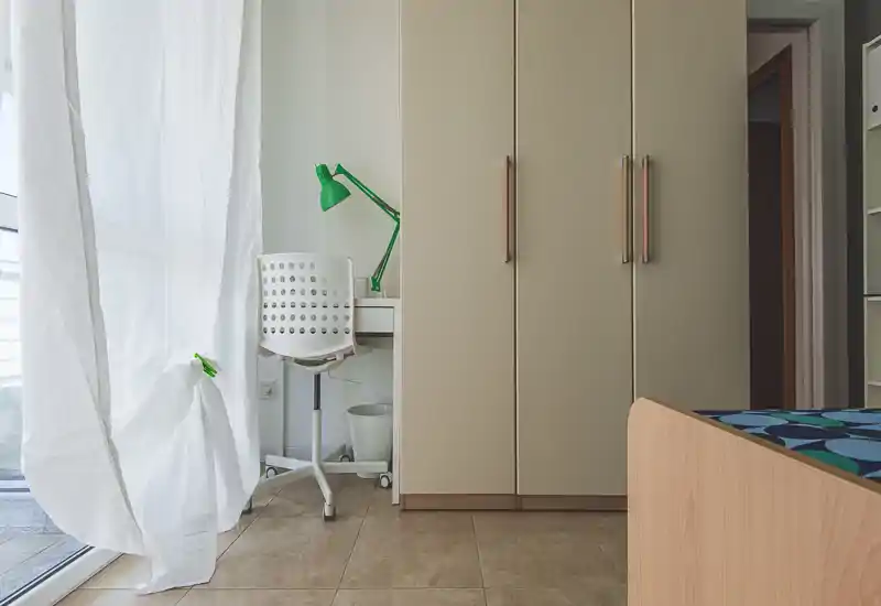 Angolo di camera luminoso e ordinato con parte del giroletto in legno, un grande armadio e una piccola scrivania con lampada verde vicino a una tenda leggera che lascia entrare luce naturale.