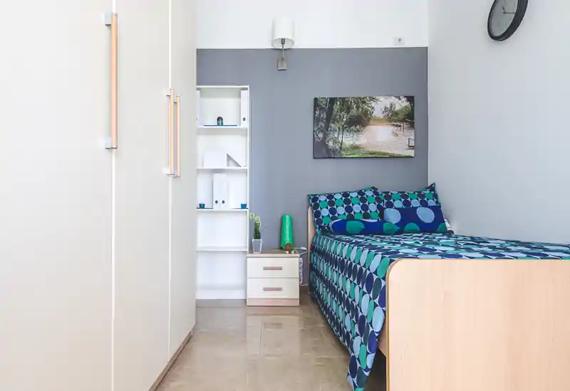 Camera singola luminosa e ordinata con biancheria a motivi turchesi, comodino e armadio a muro; stile moderno e pulito, perfetta per un annuncio.