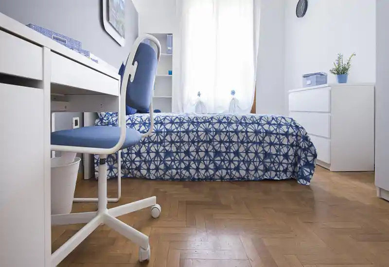 Camera singola luminosa e ordinata con copriletto blu a motivo, scrivania e sedia vicino alla finestra; parquet chiaro e mobili bianchi creano un’atmosfera fresca e ariosa.