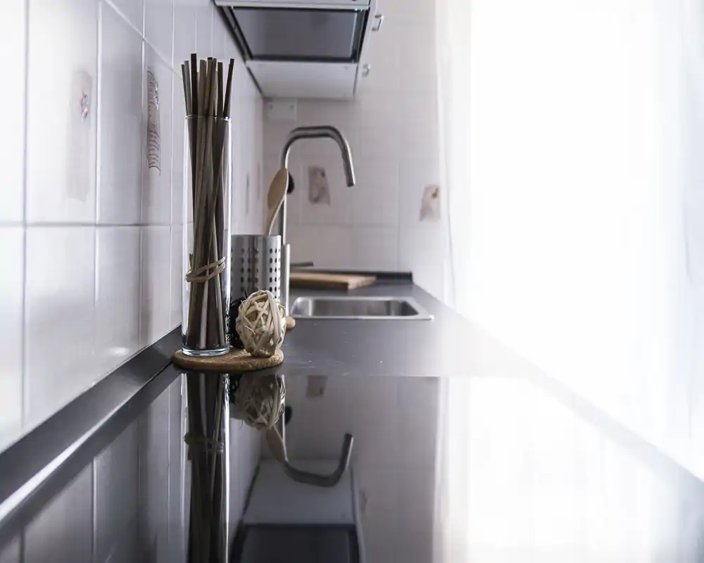 Encimera de cocina moderna y luminosa con superficie reflectante, fregadero y soporte de utensilios elegante; la luz natural suave desde la derecha crea un ambiente acogedor.
