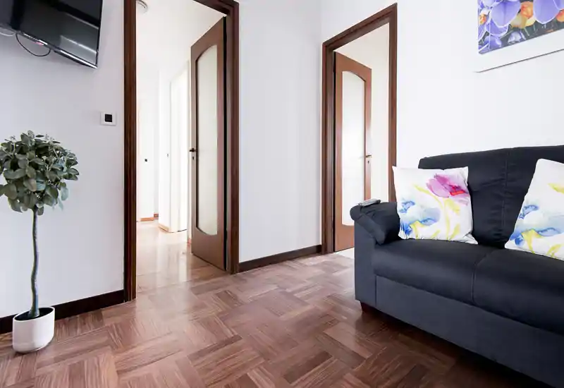 Zona living luminosa e ben arredata con divano scuro, cuscini floreali colorati, pianta in vaso e pavimento in parquet — perfetta per foto di annuncio che evidenziano comfort e decoro pulito.