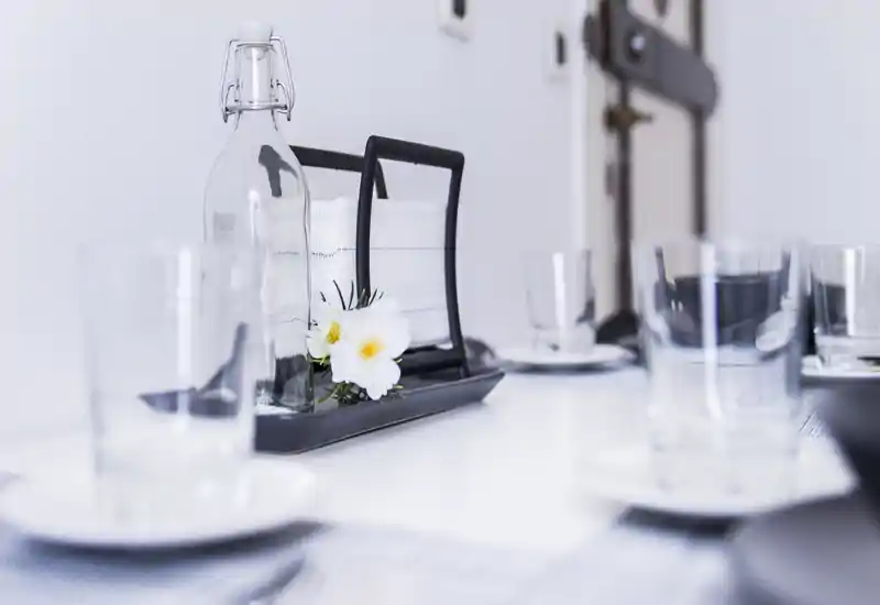 Primo piano di un tavolo da pranzo apparecchiato con bicchieri, una bottiglia d'acqua e un portatovaglioli con piccoli fiori decorativi, scattato con profondità di campo ridotta e luce soffusa e luminosa.
