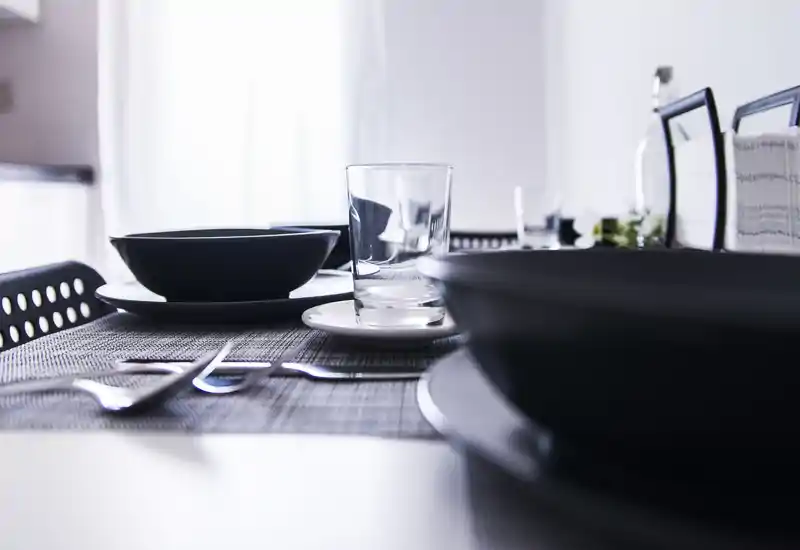 Allestimento da pranzo moderno con ciotole scure, bicchieri e posate su un sottopiatto intrecciato; stile luminoso e minimale, perfetto per appartamenti contemporanei.