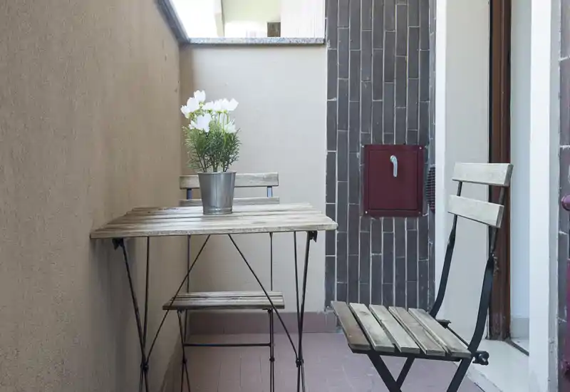 Piccolo balcone arredato con tavolino bistro in legno e due sedie pieghevoli, decorato con un vaso di fiori bianchi; angolo accogliente per il caffè.