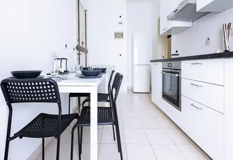Cucina moderna e luminosa con forno incassato, frigorifero e un piccolo tavolo da pranzo per quattro; mobili bianchi e sedie nere minimal creano uno spazio funzionale e pulito.