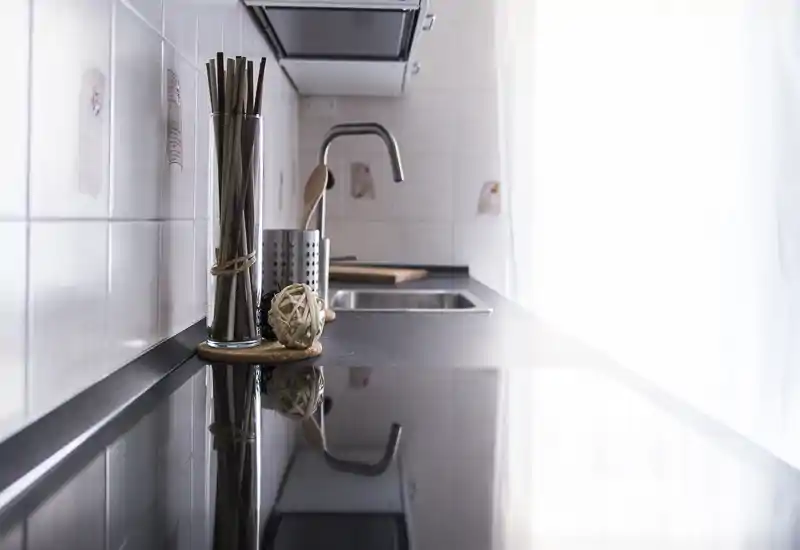 Bancone cucina moderno e luminoso con piano riflettente, lavabo e portautensili elegante; luce naturale soffusa da destra crea un’atmosfera accogliente.